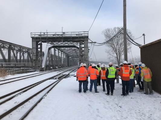 A group of people in reflective vests and hard hats stand in the snow in front of a large grey bridge outside.