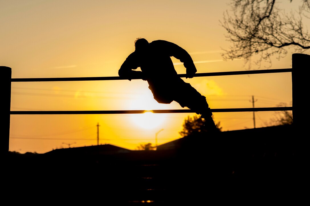 A Marine shown in silhouette climbs over an obstacle as the sun shines behind.