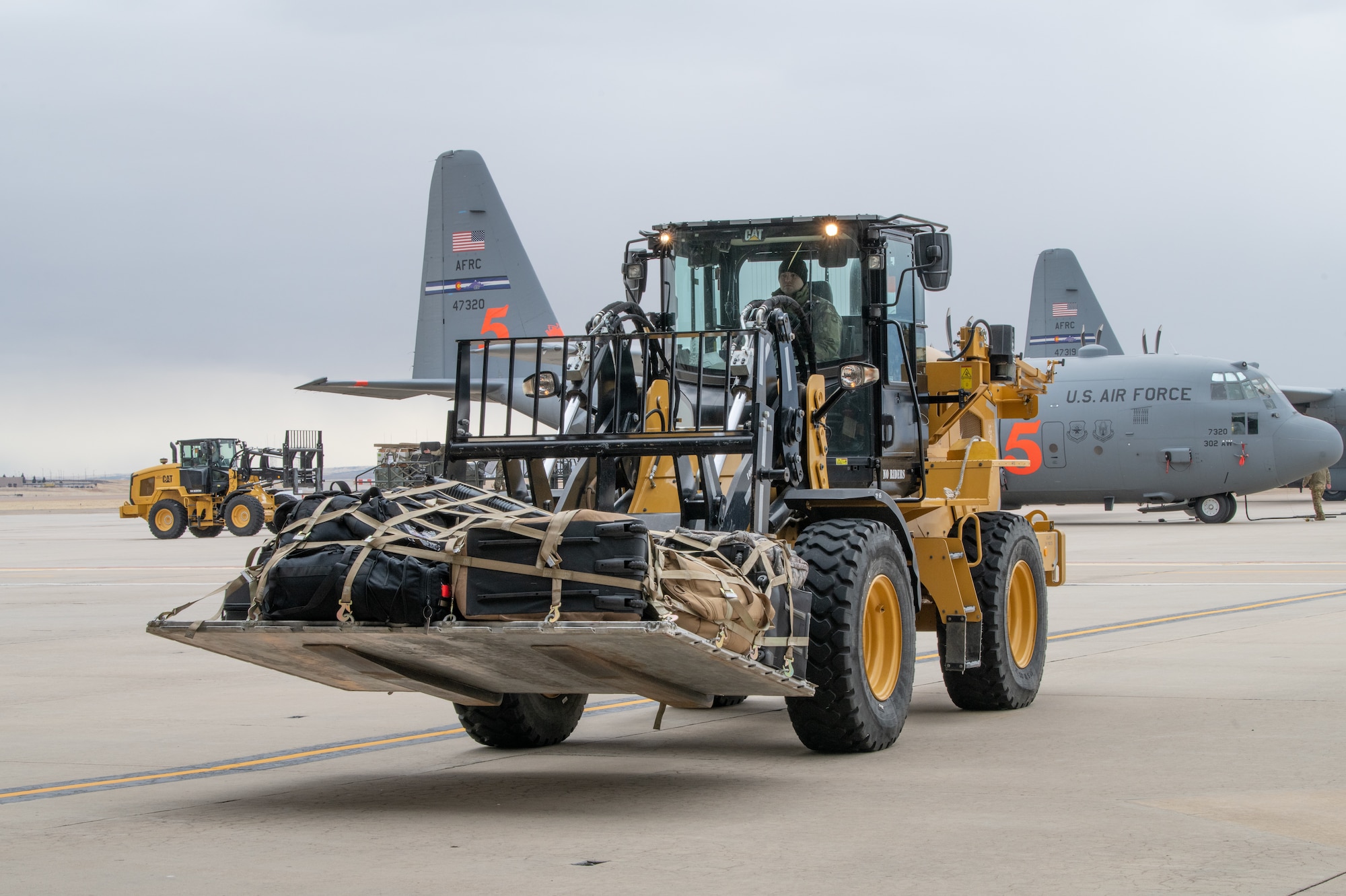 Large forklift carrying heavy pallet drives toward camera with military plane in the background