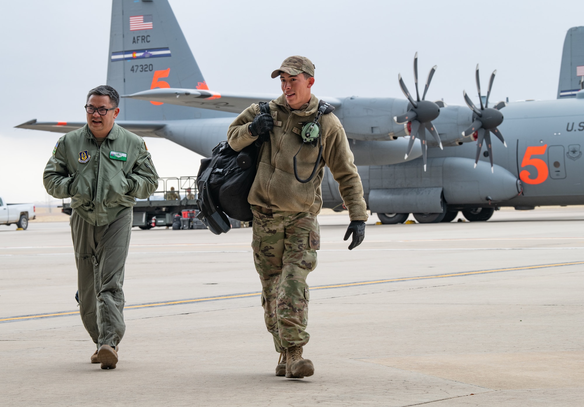 Two men in military uniform walk toward the camera with a military plane in the background