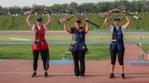 Three women in shooting uniforms posing in celebration with their shotguns outside at event.