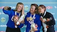 three women in their country's uniform posing for selfie with medals in front of ISSF backdrop.