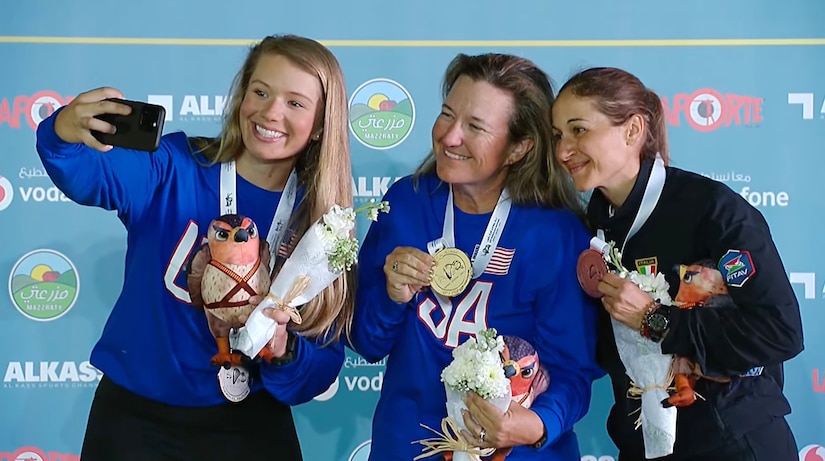 three women in their country's uniform posing for selfie with medals in front of ISSF backdrop.