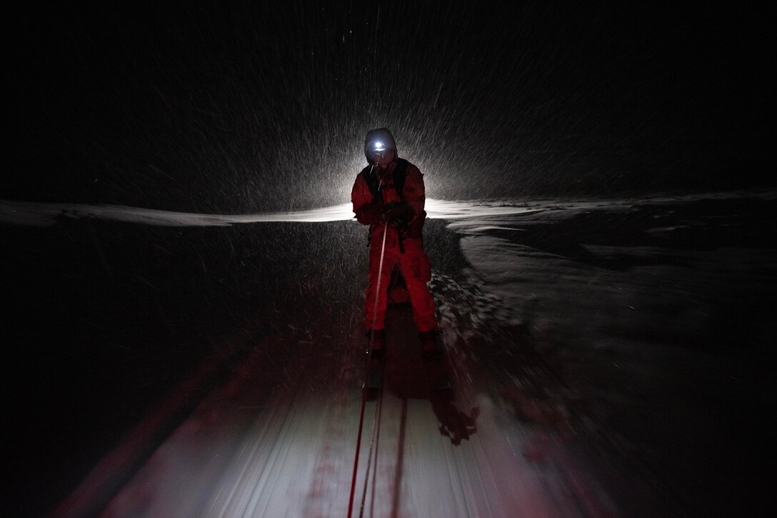 An airman wearing skis is pulled by a snow machine at night.