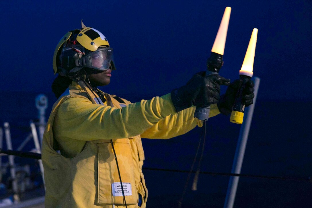 A sailor holds two flashlights at arm’s length as a signal at night.