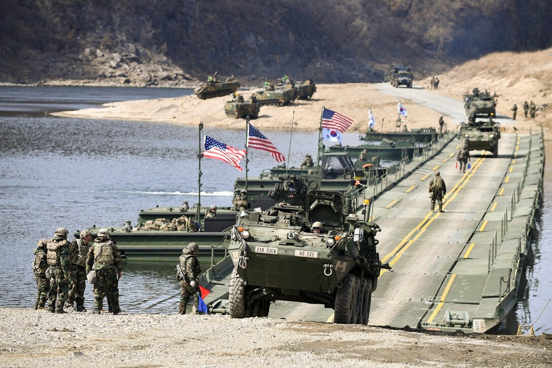 Soldiers drive vehicles over a temporary bridge as fellow soldiers watch with mountains in the background.