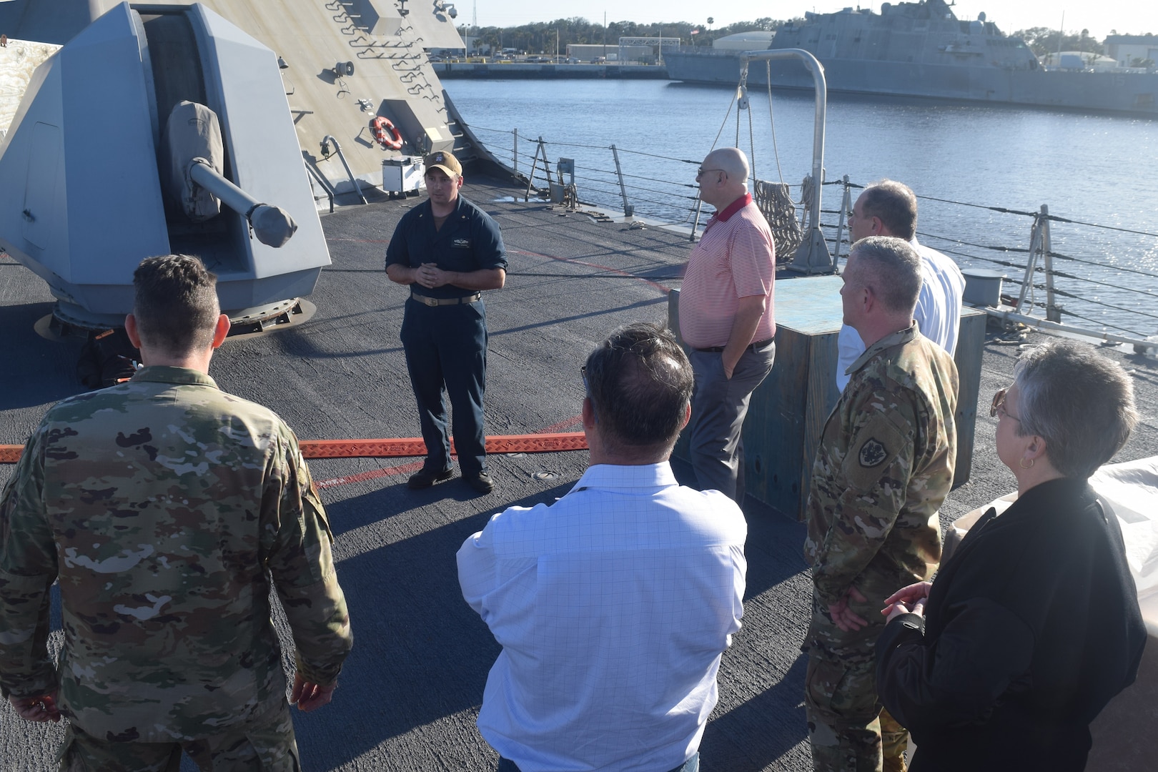 A Navy officer speaks to a small group of people on the deck of a ship.