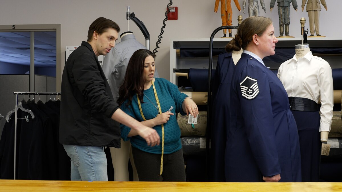 Two Air Force clothing designers on the left side measure a pregnant Airman's uniform on the right side