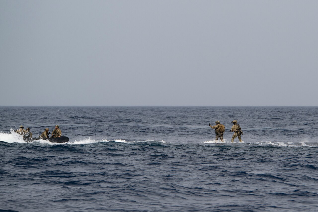 Service members sit in a rubber craft while others stand in the water,