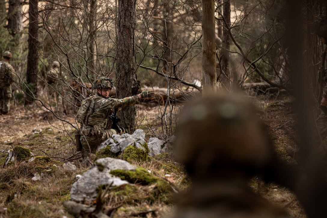 A soldier kneels and points ahead as other soldiers stand around him.