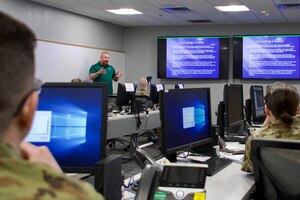 People sit at computer monitors while a man in green shirt presents information from large monitors.