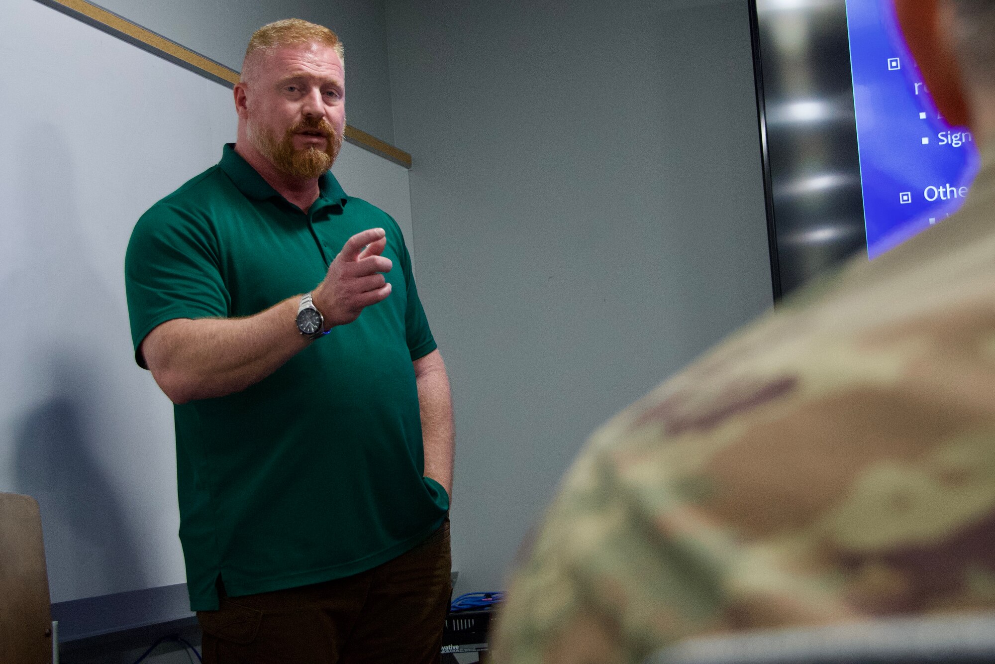 Man in green shirt holds hand up while explaining emergency preparedness to military members.