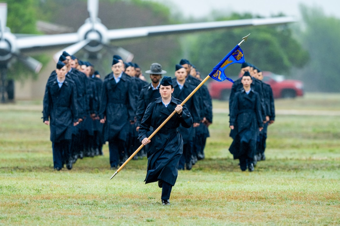 Air Force Basic Military Training graduates walk across the grass with an aircraft behind them.