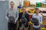 Engineers in Naval Surface Warfare Center, Carderock Division’s Expeditionary and Development Power and Energy Branch from left: Matthew Huffman, technical area lead, Kevin Lin, electrical engineer and James Mulford, mechanical engineer, display their invention, the Lithium 6T Top Cap, in West Bethesda, Md., on March 8. The engineers are shown with three of their four prototypes, which is a modular, field-reconfigurable direct current power distribution device that can be installed and mounted on top of a Li6T battery. (U.S. Navy photo by Aaron Thomas)