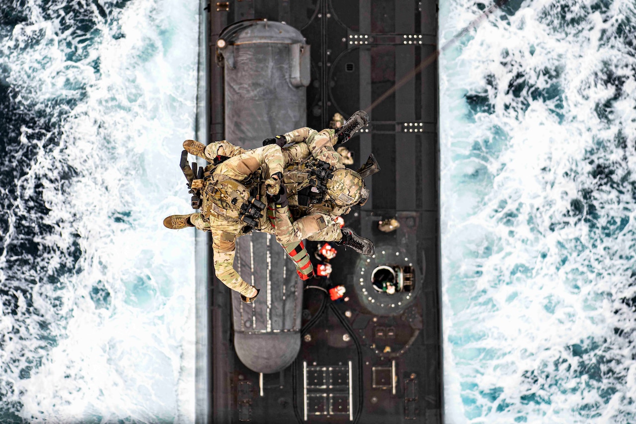 A look from above as two Navy SEALs hang from a rope over a submarine traveling through waters.