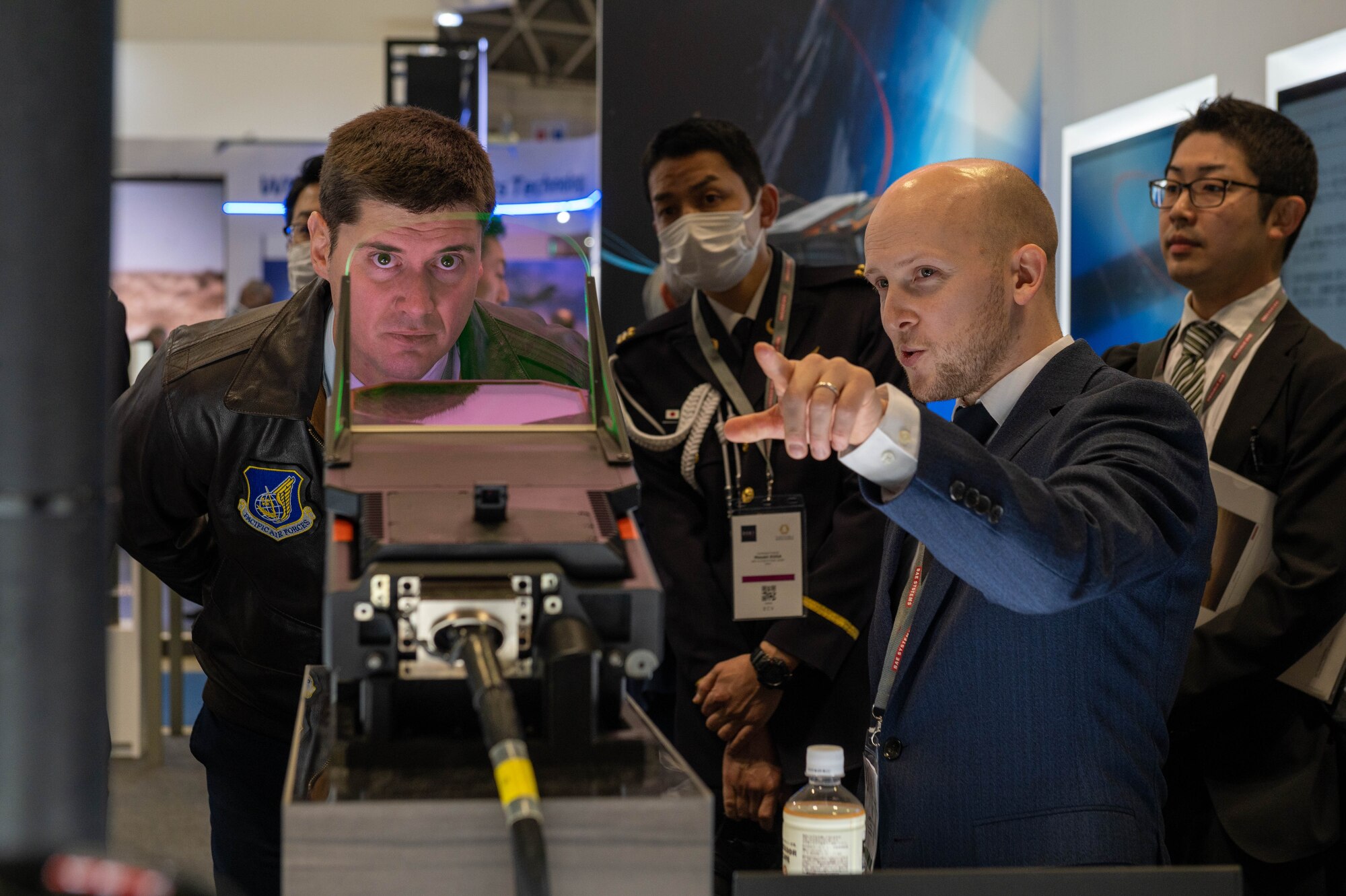 A white man wearing a leather jacket looks at a head-up display while listening to a vendor in a large convention center.
