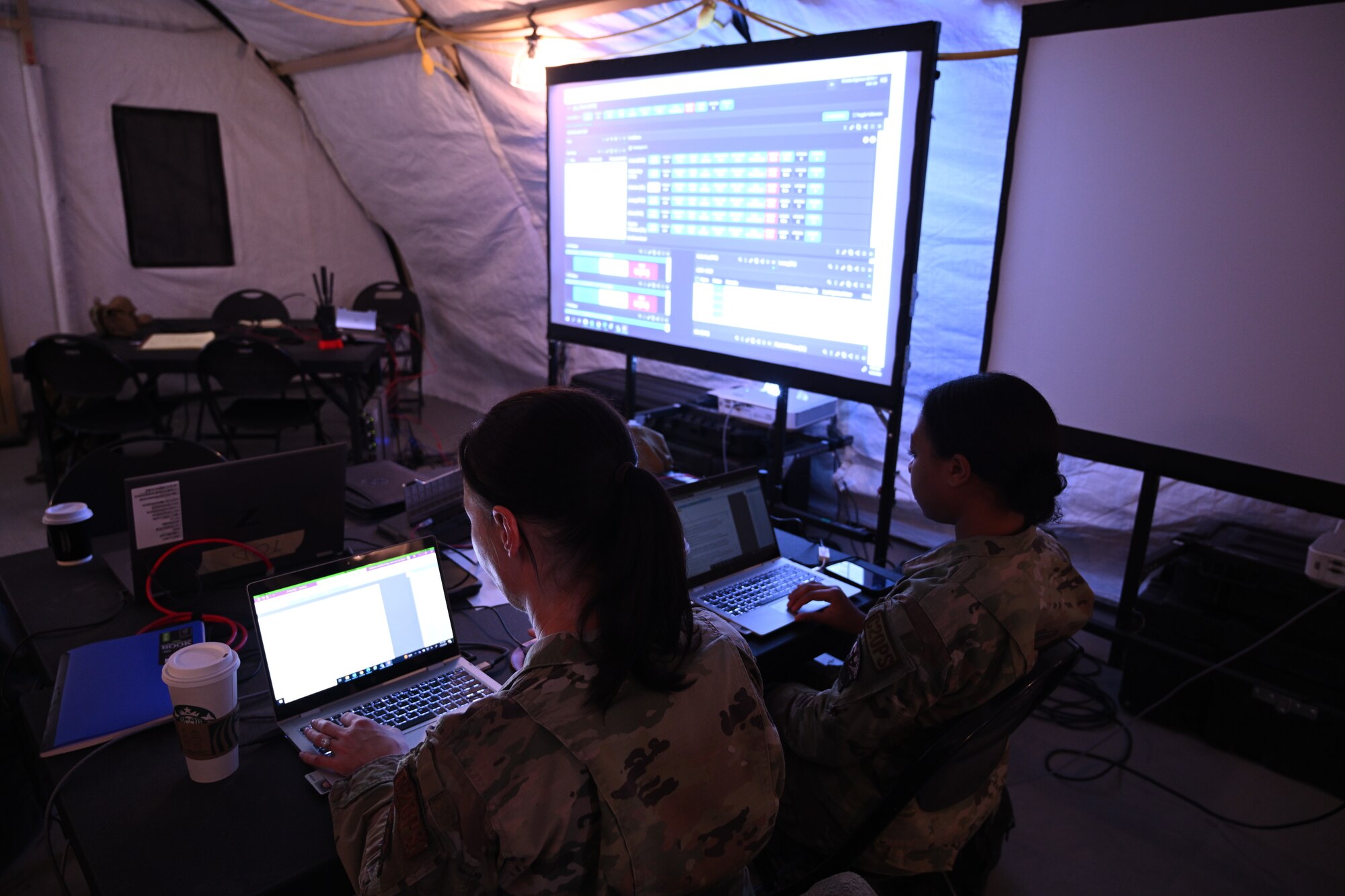 photo inside dark tent two uniformed U.S. Air Force personnel sit at computers working, in the background are two large projector screens in the background