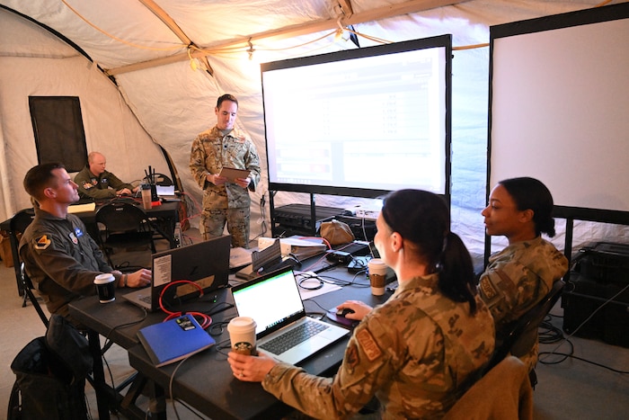 photo of four uniformed U.S. Air Force military personnel in a tent, one standing, three sitting at tables with computers with two large projector screens in the background