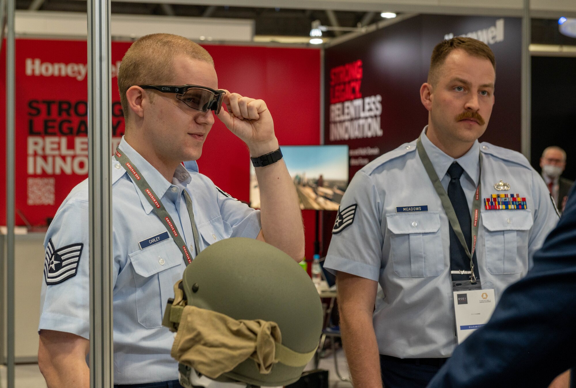 Two white men stand next to each other while listening to a vendor in a large convention center.