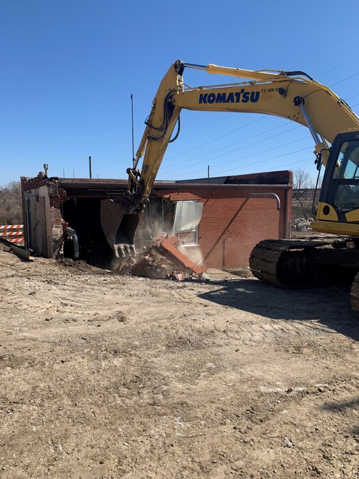 A large piece of heavy equipment demolishes a red brick building on a sunny day with blue skies in a dirt lot.