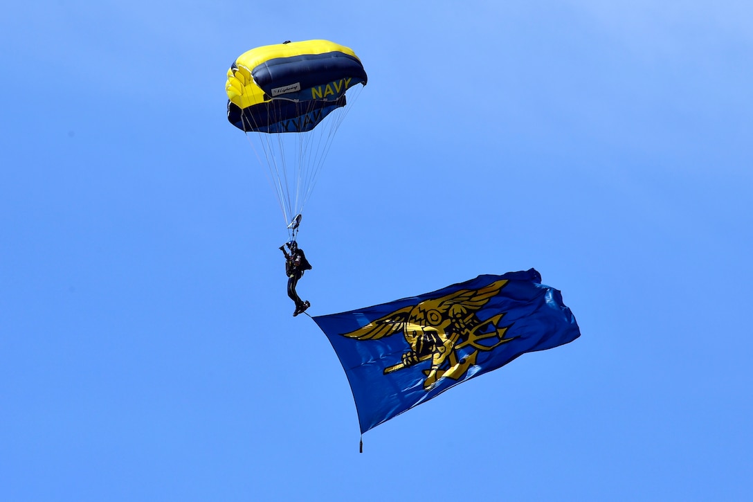 A sailor wearing a parachute displays a flag while free-falling.