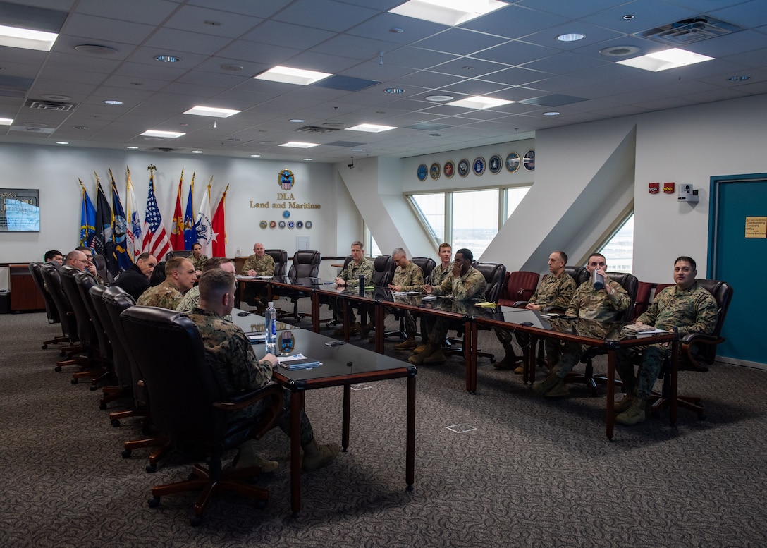 A group of people sit around a large conference room table and are all wearing military uniforms, mostly camo OCPs. They are deep in discussion.