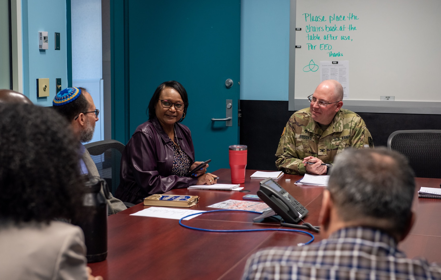 A group of people, dark skinned and light skinned sit around a table and speak to a bald man in glasses in a OCP Army uniform.