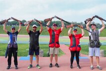Men and women in shooting uniforms posing in celebration with their shotguns outside at event.