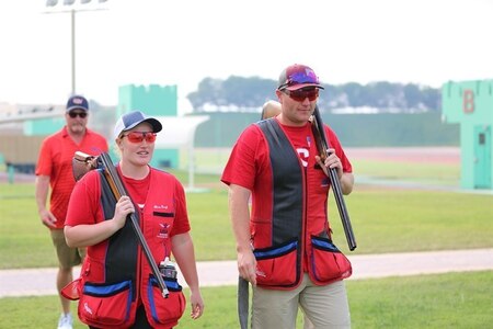 Man and woman in USA shooting uniforms walking with their shotguns outside at event.