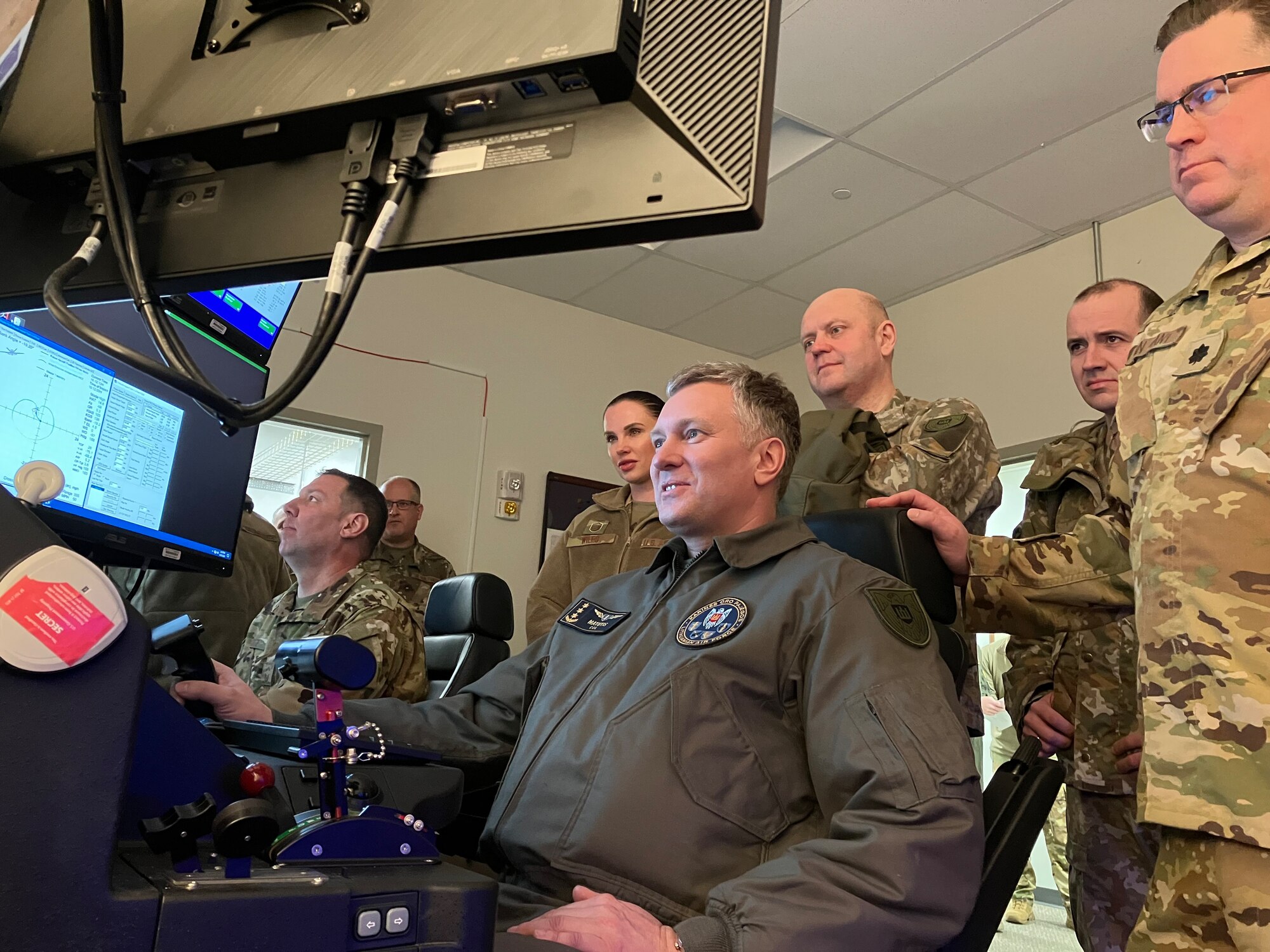 A man in a military uniform sits in a flight simulator surrounded by military personnel.