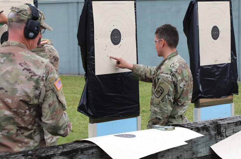 Multiple men in U.S. Army uniforms inspect targets on outdoor pistol range.