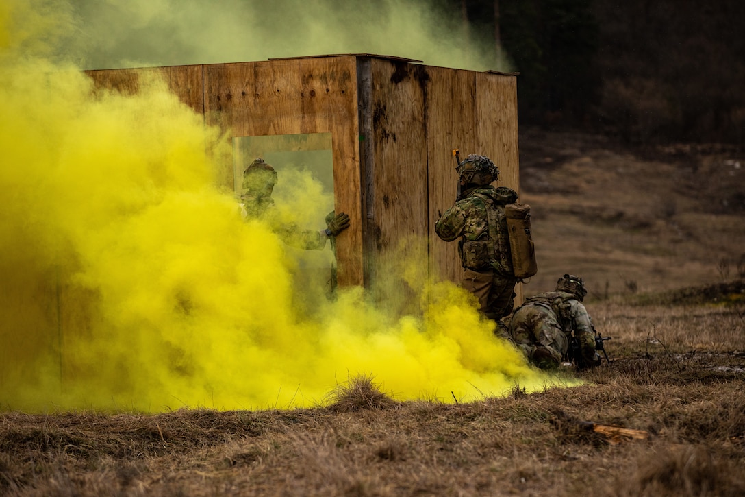 Soldiers gather around a small roofless building in a large field with yellow smoke.