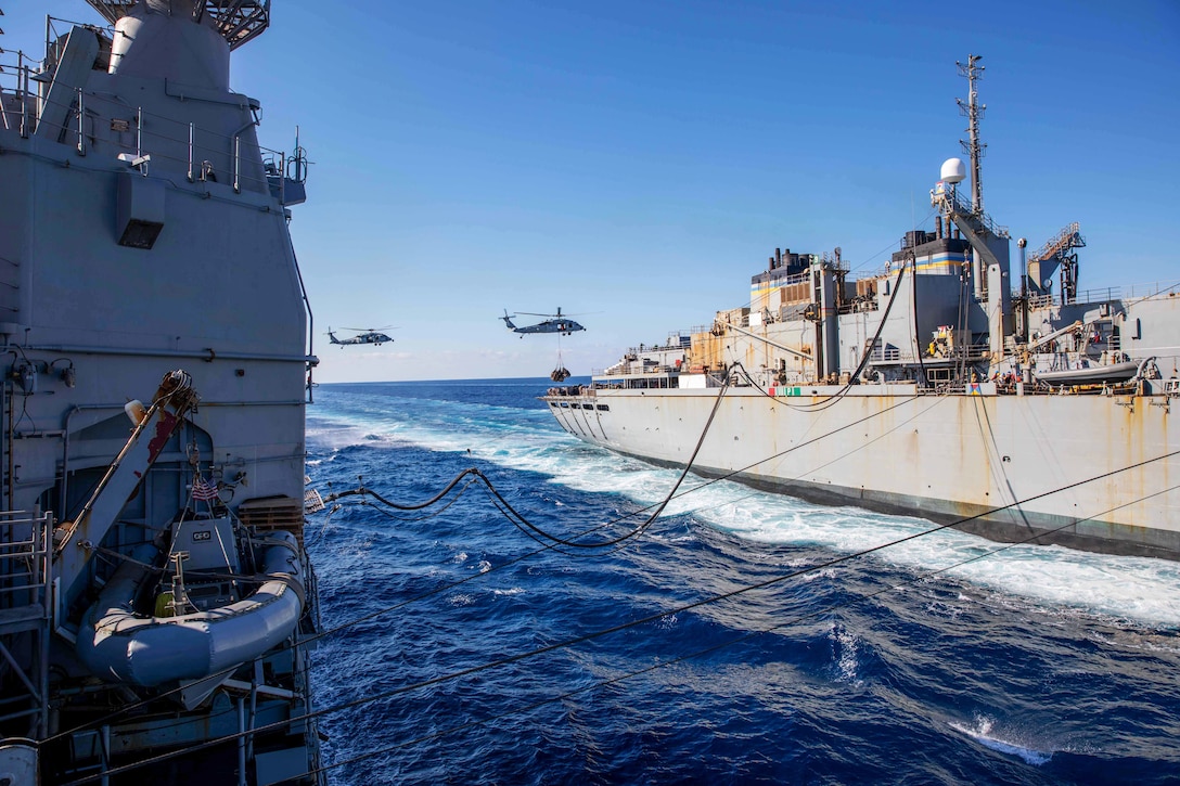 A ship performs a replenishment while two helicopters fly by.