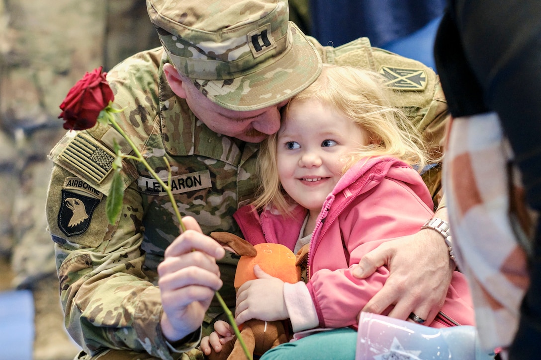 A soldier kneels hugging a child while holding a rose.