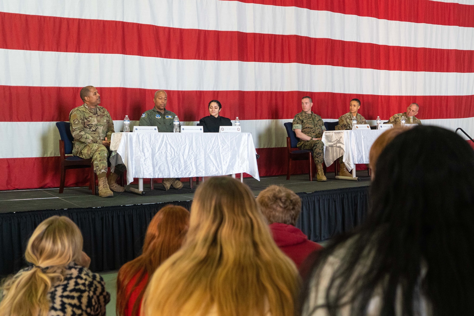 a group of military members sit on a stage in front of a flag