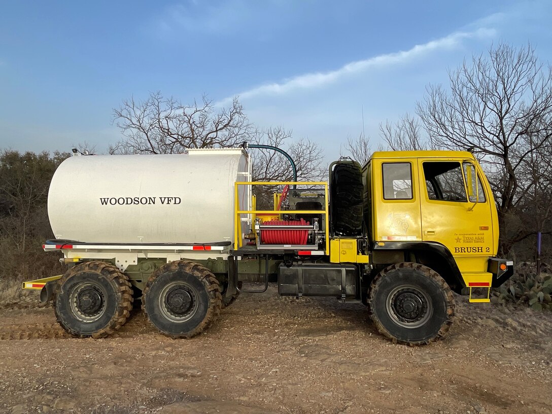 A former military supply truck sits on a patch of dry dirt patch with scrubby small trees and other brush in the background. The cab of the truck is now yellow and the back of the truck is dominated by a white water tank.