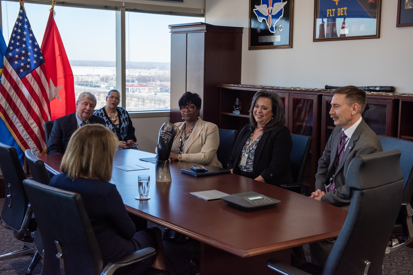 A brown haired woman in a blue dress is seated at a conference table with two men in business suits and three dark skinned and haired women in business dress. Their are flags in the background.