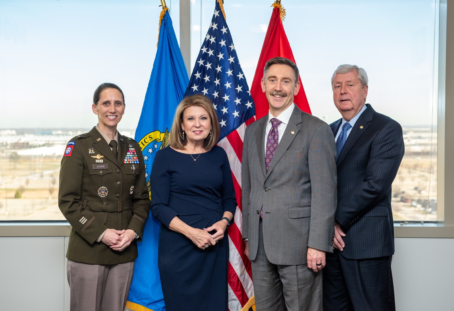 A dark haired woman in a Army dress uniform with a olive green jacket and tan pants, a brown haired woman wearing a dark blue wrap around dress, a brown haired man in a gray suit and a white haired man in a black suit pose for a photo in front of several flags in front of a window.