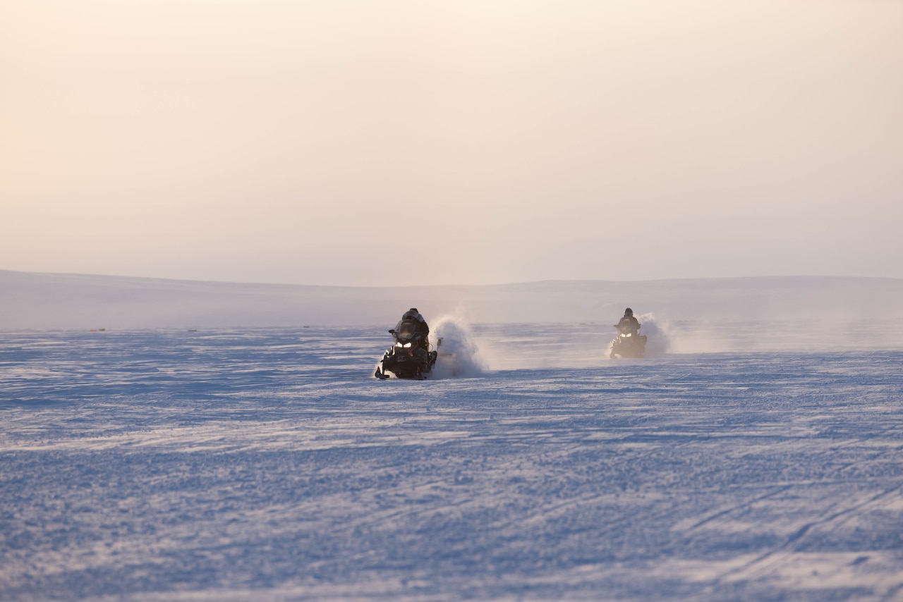 Two airmen drive snow machines across a snowy area.