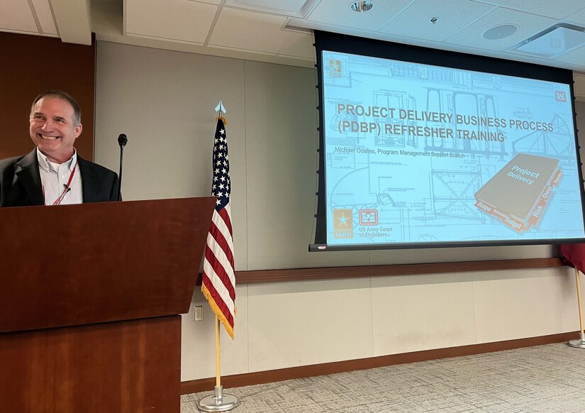 A man stands behind a podium and presents a training which is displayed on a projector screen in the background.