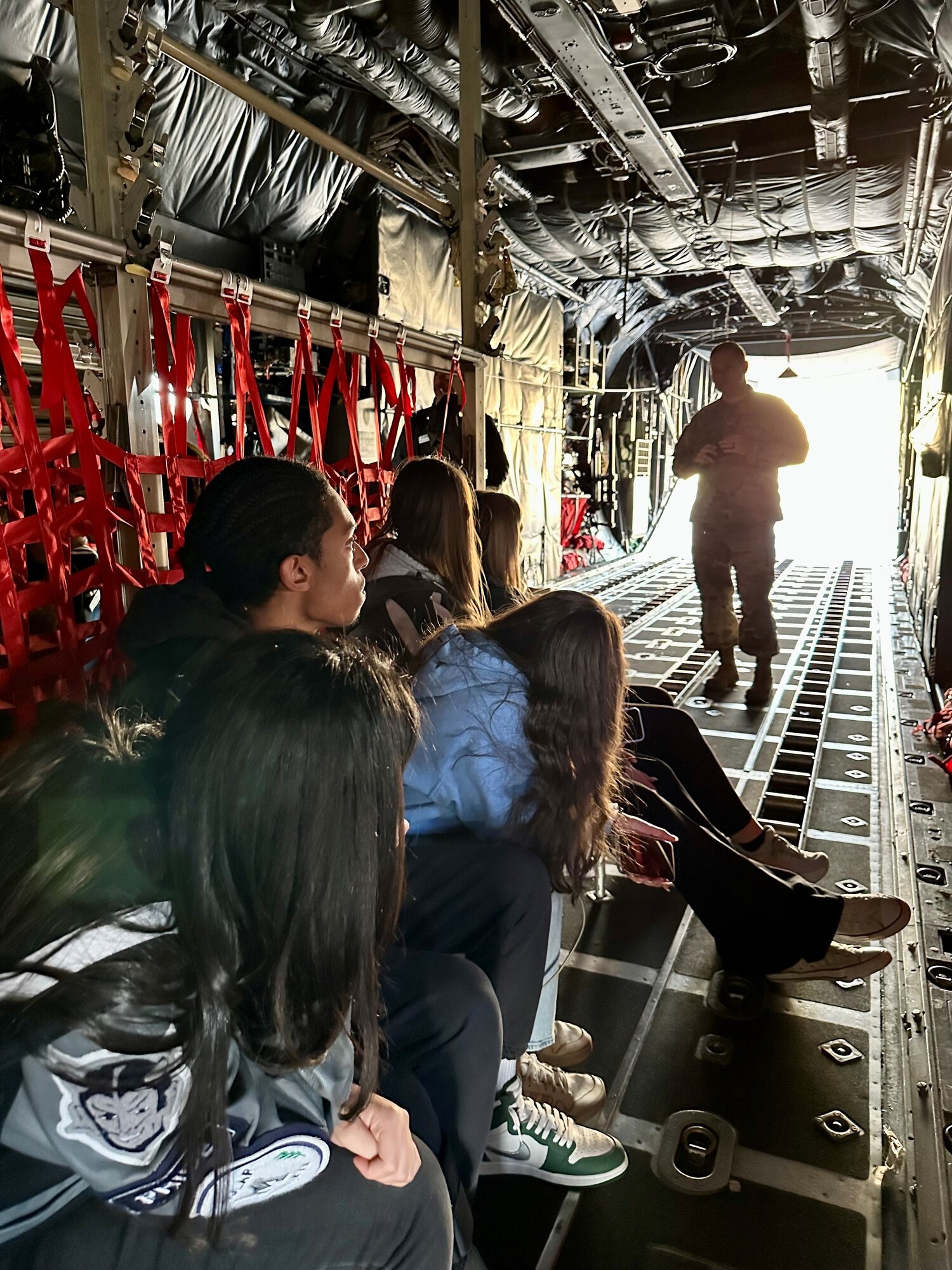 a group of people sit inside an aircraft as a military member speaks to them