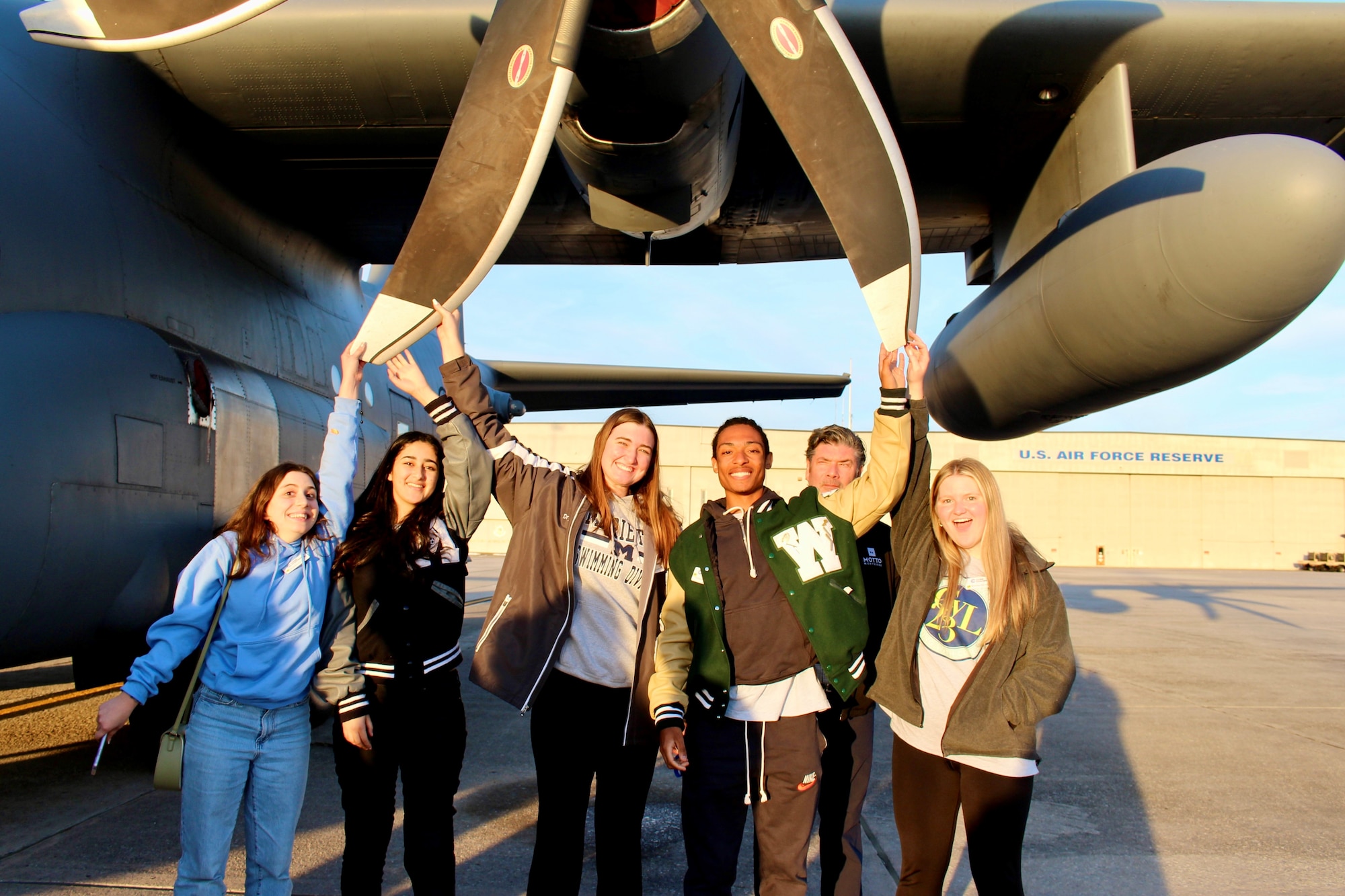a group of students touch a propeller while standing under the wing of a military aircraft