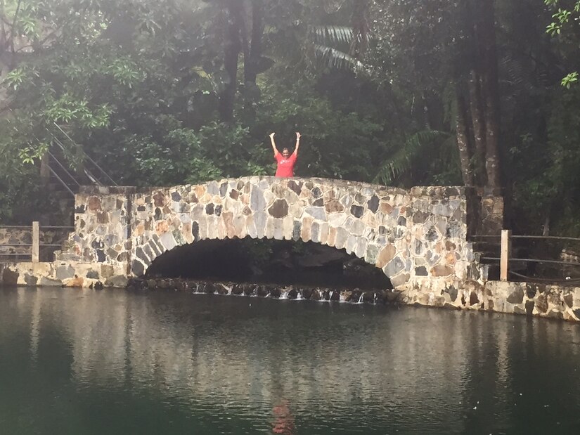 A woman stands on a bridge with her arms in the air with water in the foreground and trees in the background.