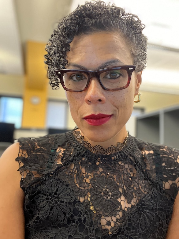 A woman sits at a desk and smiles while wearing glasses.