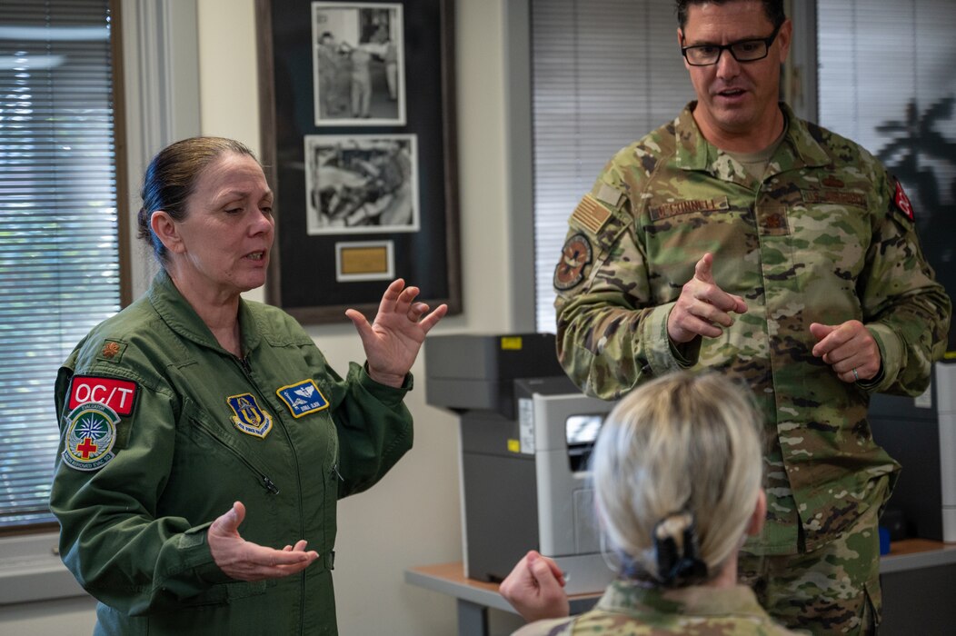 A female Airman in her flight suit instructs two other airmen as they stand around a table in an office.