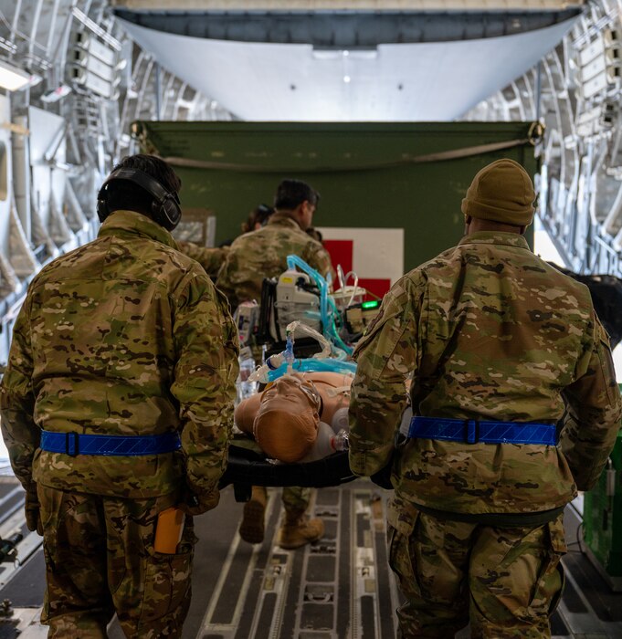 Three Airmen carry a stretcher with a training medical manikin out of a C-17 Globemaster where there is a green medical supply box in the back.