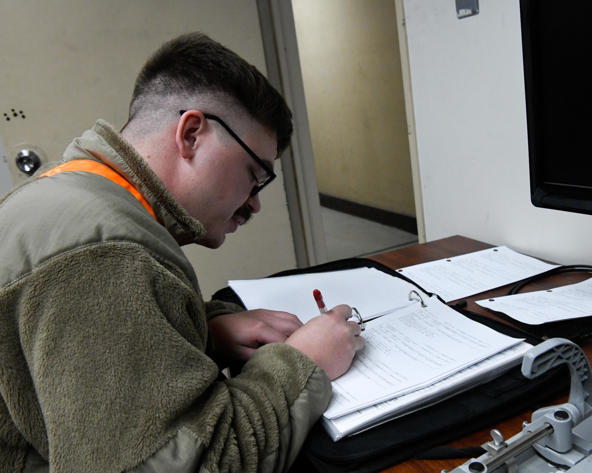 A white man wearing a green fleece jacket sits at a desk, writing on some paper.