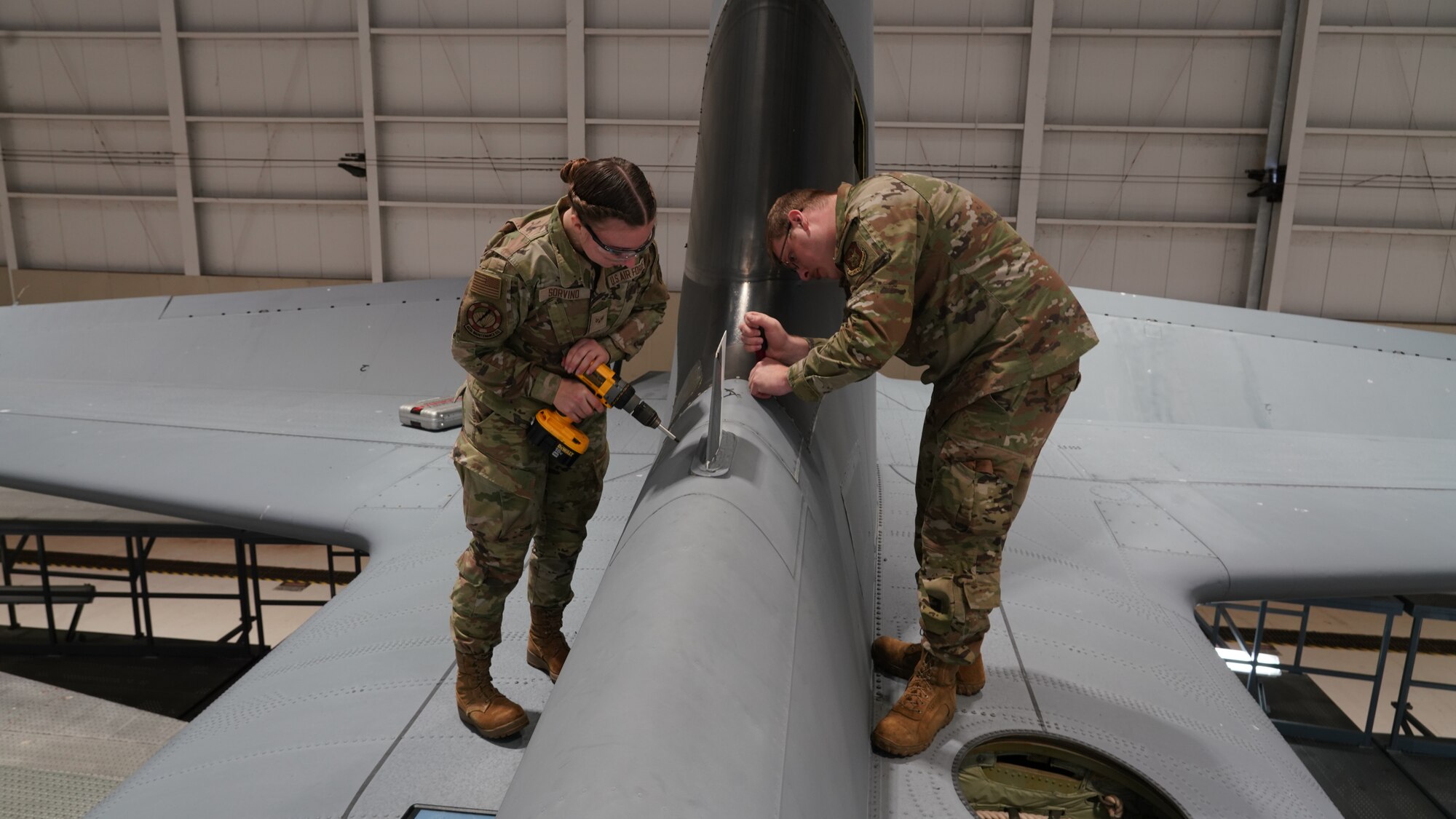 Female Airman removes screws from panel while male Airman removes sealant from panel.
