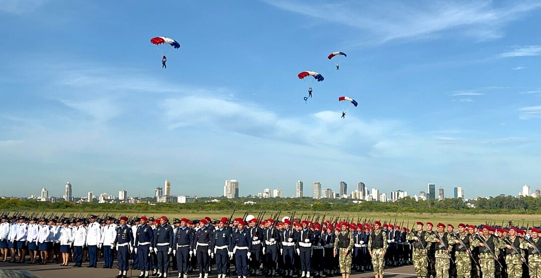 Military members parachute into the Central Ceremony of the Centennial Paraguayan Military Aviation while military members stand at attention in Asunción, Paraguay, Feb. 22, 2023. Senior leaders from 10 countries attended the event to honor Paraguayan Air Force’s Centennial Anniversary. (Courtesy photo)