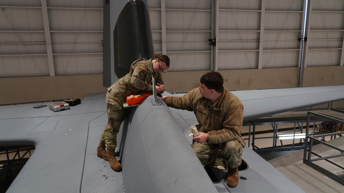 Female Airman removes part from aircraft with male Airman holding the part steady during removal.